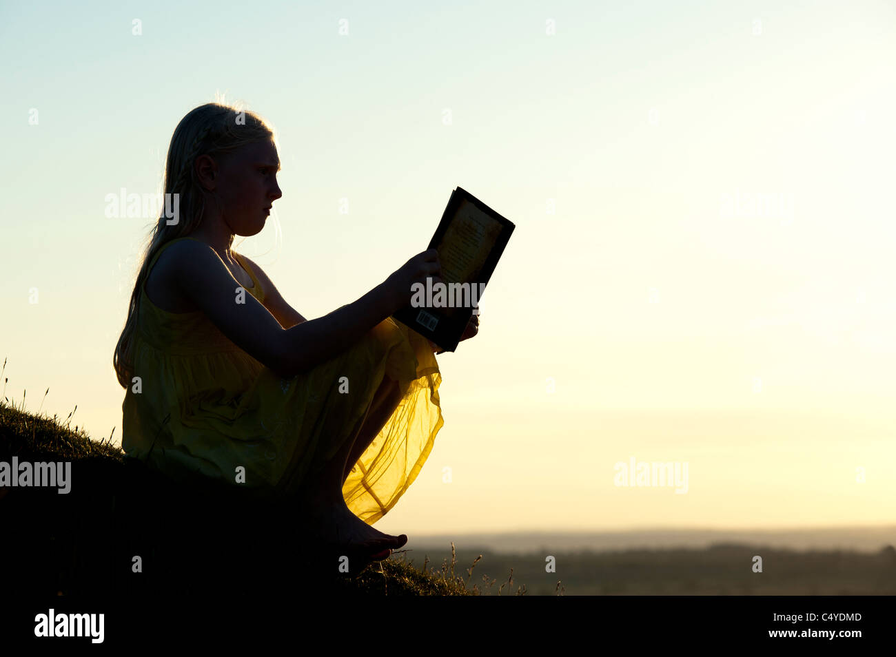 Junges Mädchen sitzt auf einem Hügel bei Sonnenuntergang ein Buch zu lesen. Silhouette Stockfoto