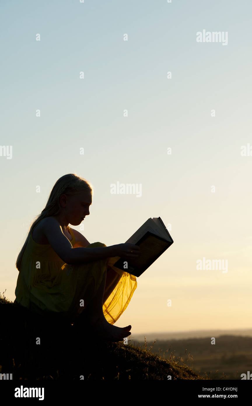 Junges Mädchen sitzt auf einem Hügel bei Sonnenuntergang ein Buch zu lesen. Silhouette Stockfoto