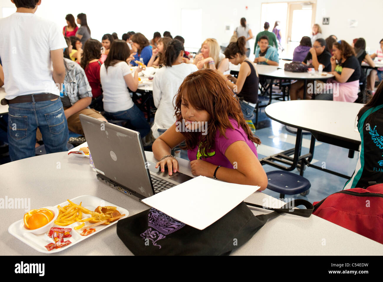 Hispanische Teen Student arbeitet auf Laptop-Computer in der Cafeteria der Schule, während andere mit Freunden sozialisieren und zu Mittag essen Stockfoto