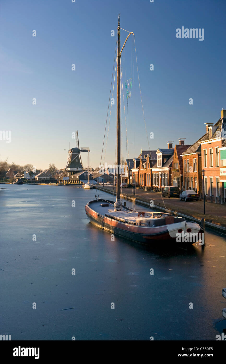 Die Niederlande, Woudsend, traditionelle Frachtschiff, jetzt Segelboot für den Tourismus in gefrorenen Kanal. Hintergrund Windmühle. Stockfoto