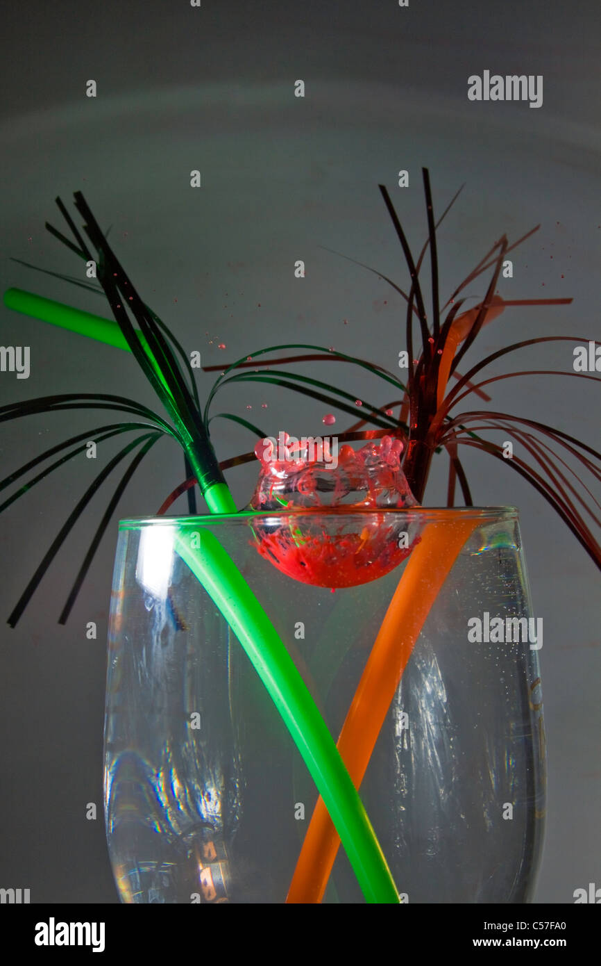 ein Tropfen rote Lebensmittelfarbe, die Kollision mit einem Glas Wasser, mit einem roten und grünen Stroh in Stockfoto