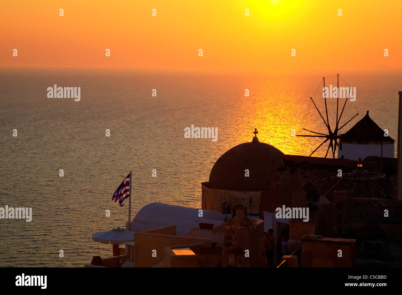 Sonnenuntergang in Oia, legendäre griechische Stadt auf der Insel Santorin, Orange Leuchten und Reflexion. Windmühle und griechische Flagge in der Abenddämmerung Stockfoto