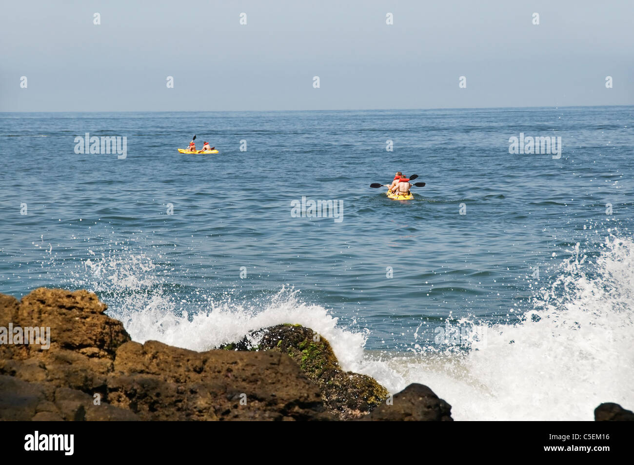 Wellen brechen sich an der felsigen Küste im Vordergrund während zwei Teams der Kajakfahrer begeben Sie sich in die Bucht von Banderas. Stockfoto