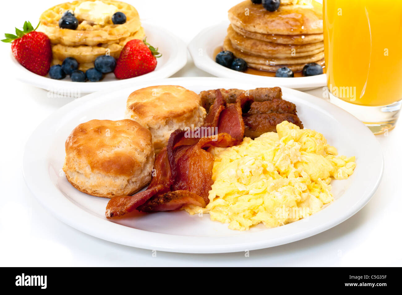 Frühstücksteller mit Rührei, Speck und Kekse Buttermilch.  Waffeln, Pfannkuchen und Orangensaft im Hintergrund. Stockfoto