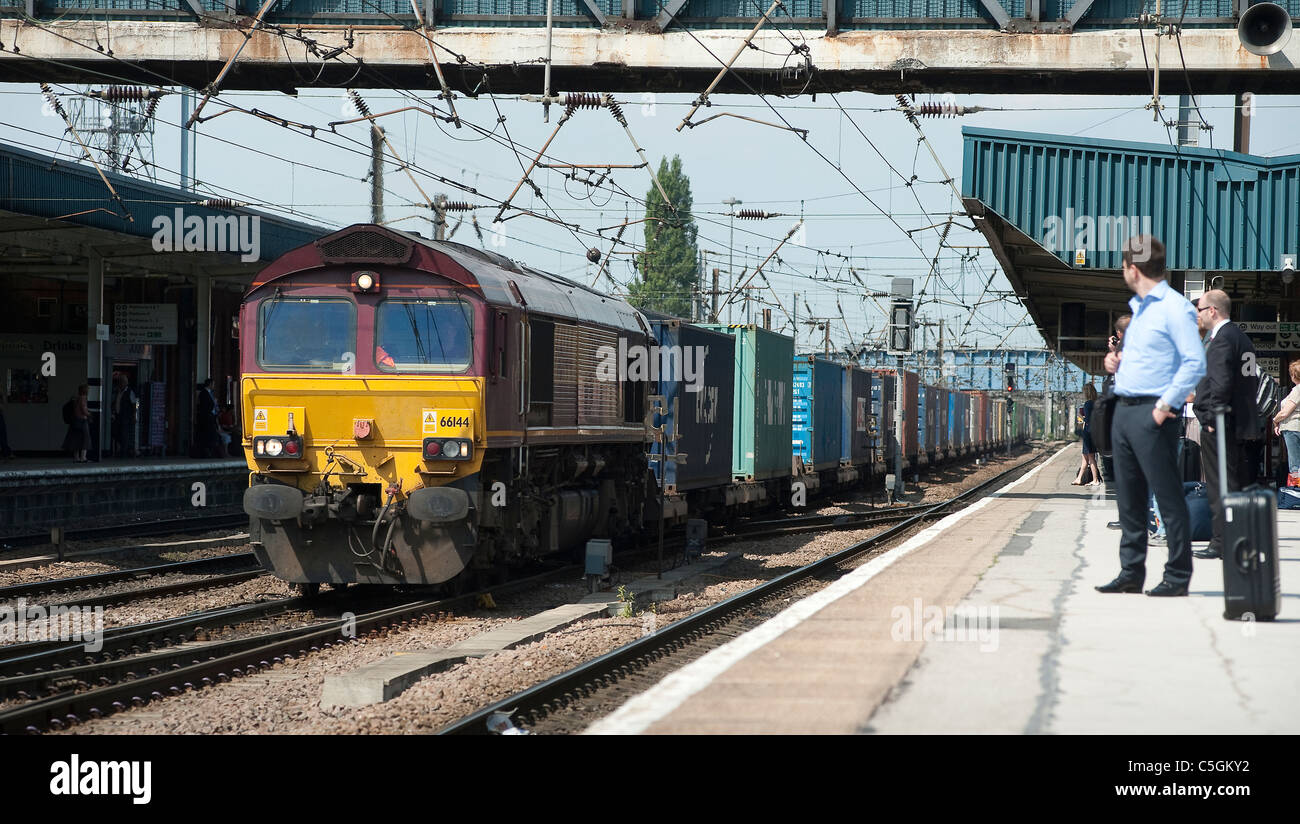 Passagiere warten auf eine Plattform als ein ein Güterzug durchläuft einen Bahnhof in England. Stockfoto