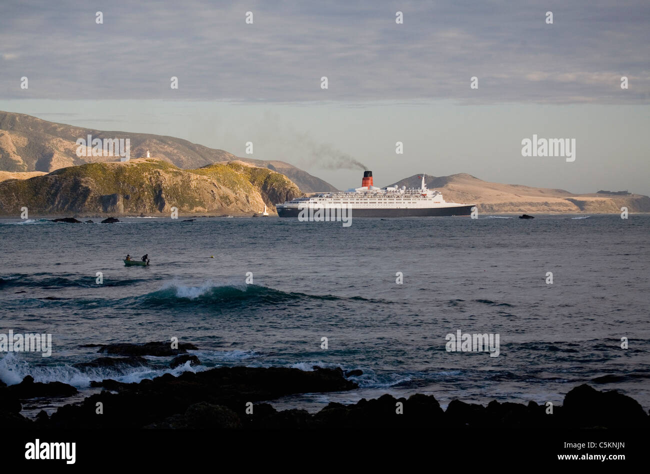 Kreuzfahrtschiff Queen Elizabeth 2 Segeln vorbei an Hügeln am Eingang nach Wellington Harbour, New Zealand, zwei Männer, die aus einer kleinen Fischen Stockfoto