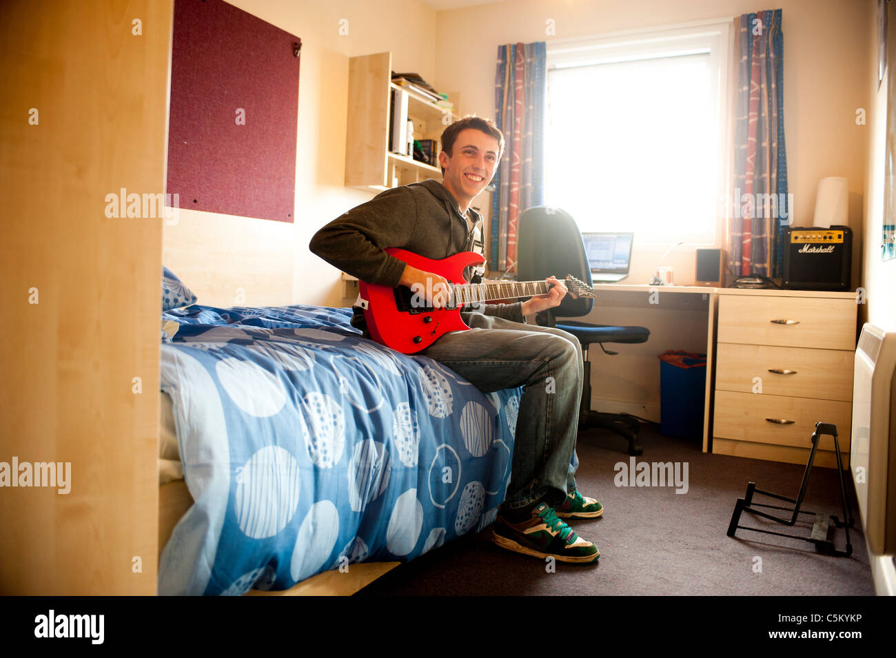 Junger kaukasischer Student, der in seinem Zimmer in den Studentenwohnheimen auf seinem Bett sitzt und eine rote Gitarre spielt. Stockfoto Junger kaukasischer Student, der in seinem Zimmer in den Studentenwohnheimen auf seinem Bett sitzt und eine rote Gitarre spielt. Stockfoto