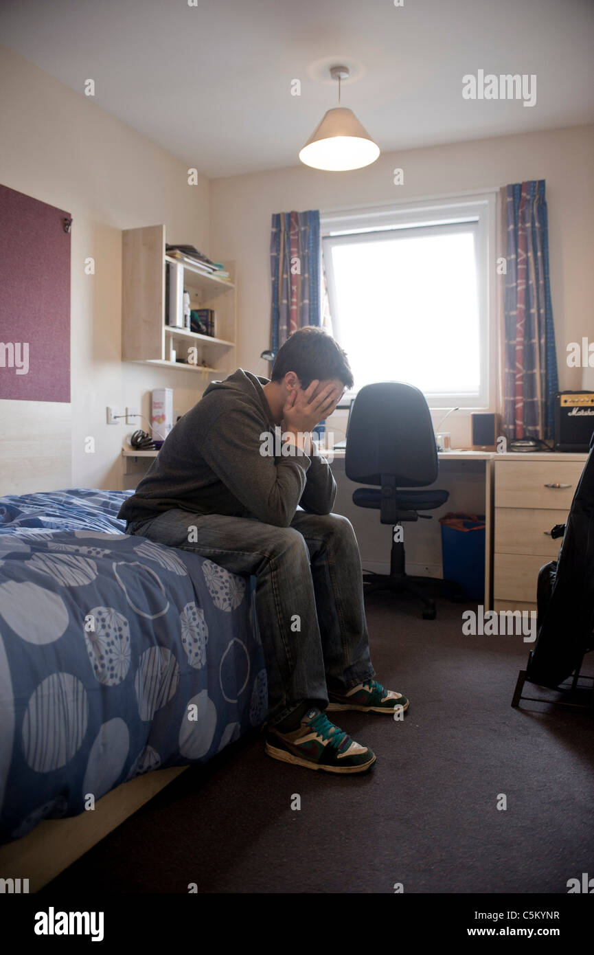 Junger kaukasischer Student. Kopf in den Händen, auf einem Bett sitzend, allein im Zimmer weg von zu Hause - Heimweh. Stockfoto