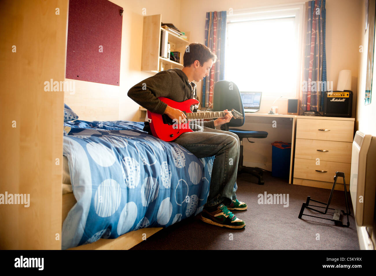 Junger kaukasischer Student, der in seinem Zimmer in den Studentenwohnheimen auf seinem Bett sitzt und eine rote Gitarre spielt. Stockfoto Junger kaukasischer Student, der in seinem Zimmer in den Studentenwohnheimen auf seinem Bett sitzt und eine rote Gitarre spielt. Stockfoto