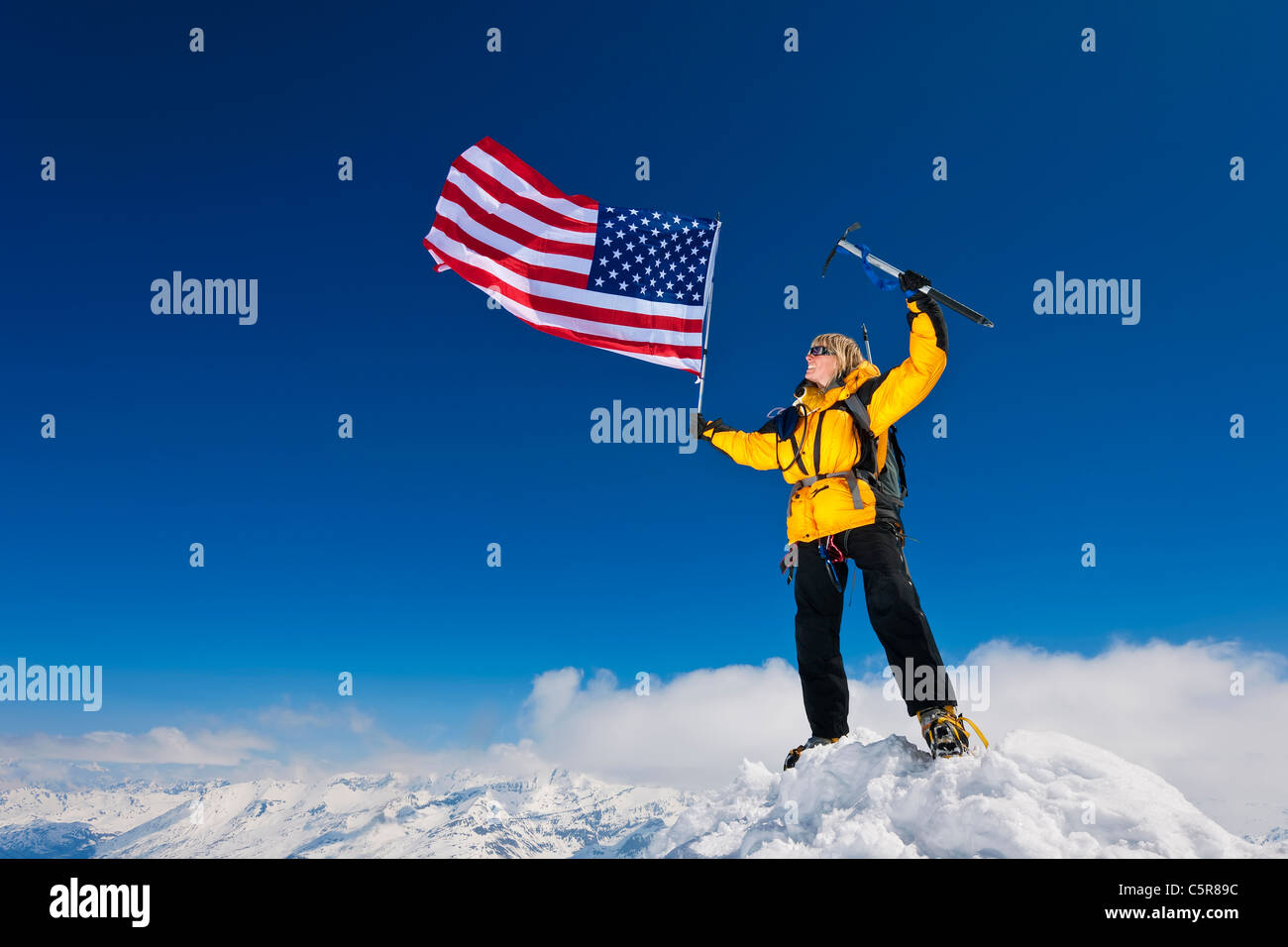 Weibliche Bergsteiger fliegen die Sterne und Streifen auf dem Gipfel des schneebedeckten Berge. Stockfoto