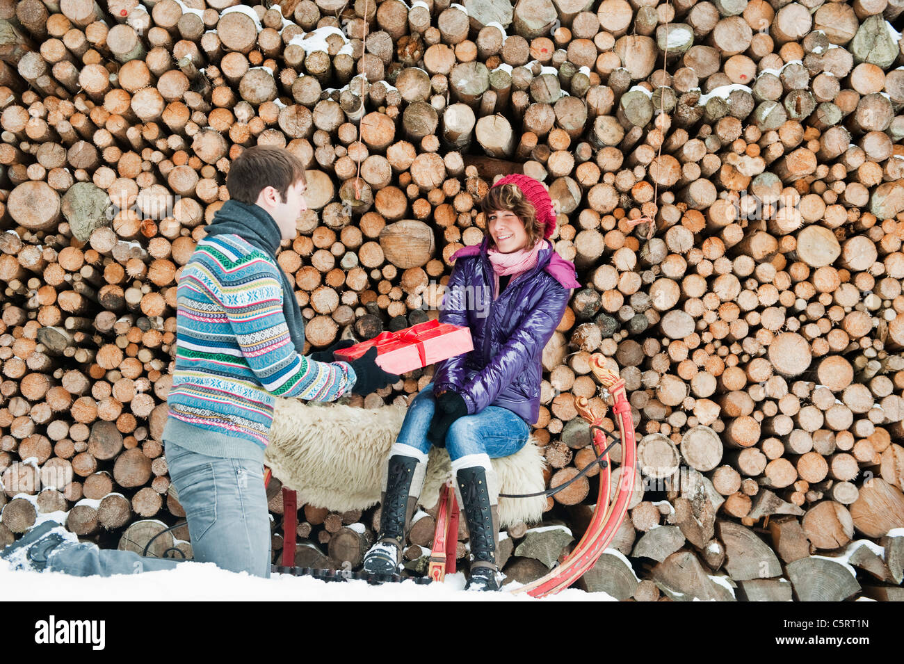 Österreich, Salzburger Land, Flachau, junger Mann geben Weihnachtsgeschenk für Frau sitzt auf Schlitten mit Brennhölzer im Hintergrund Stockfoto