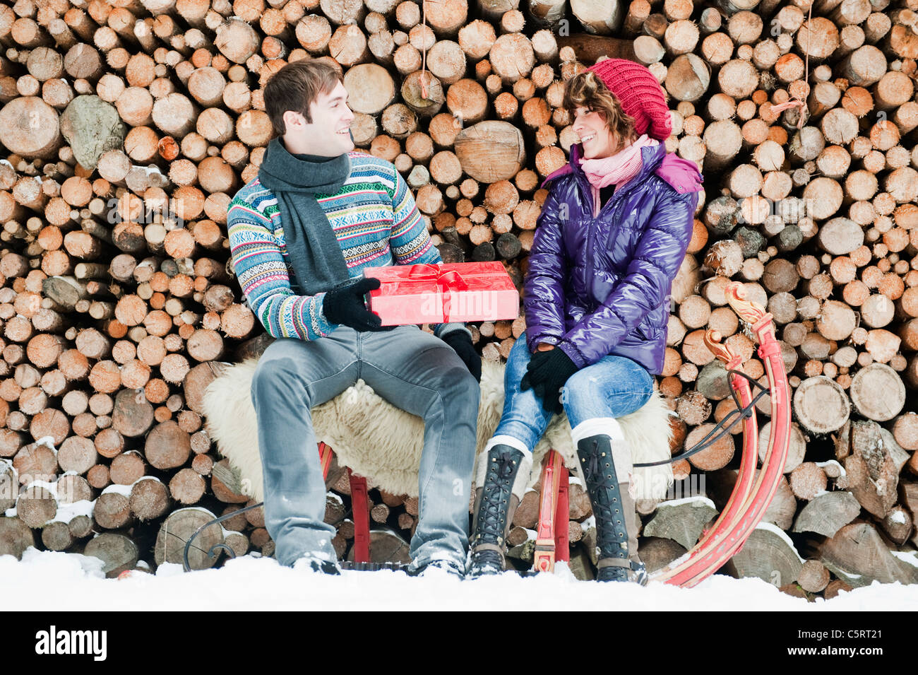 Österreich, Salzburger Land, Flachau, junger Mann geben Weihnachtsgeschenk für Frau sitzt auf Schlitten mit Brennhölzer im Hintergrund Stockfoto