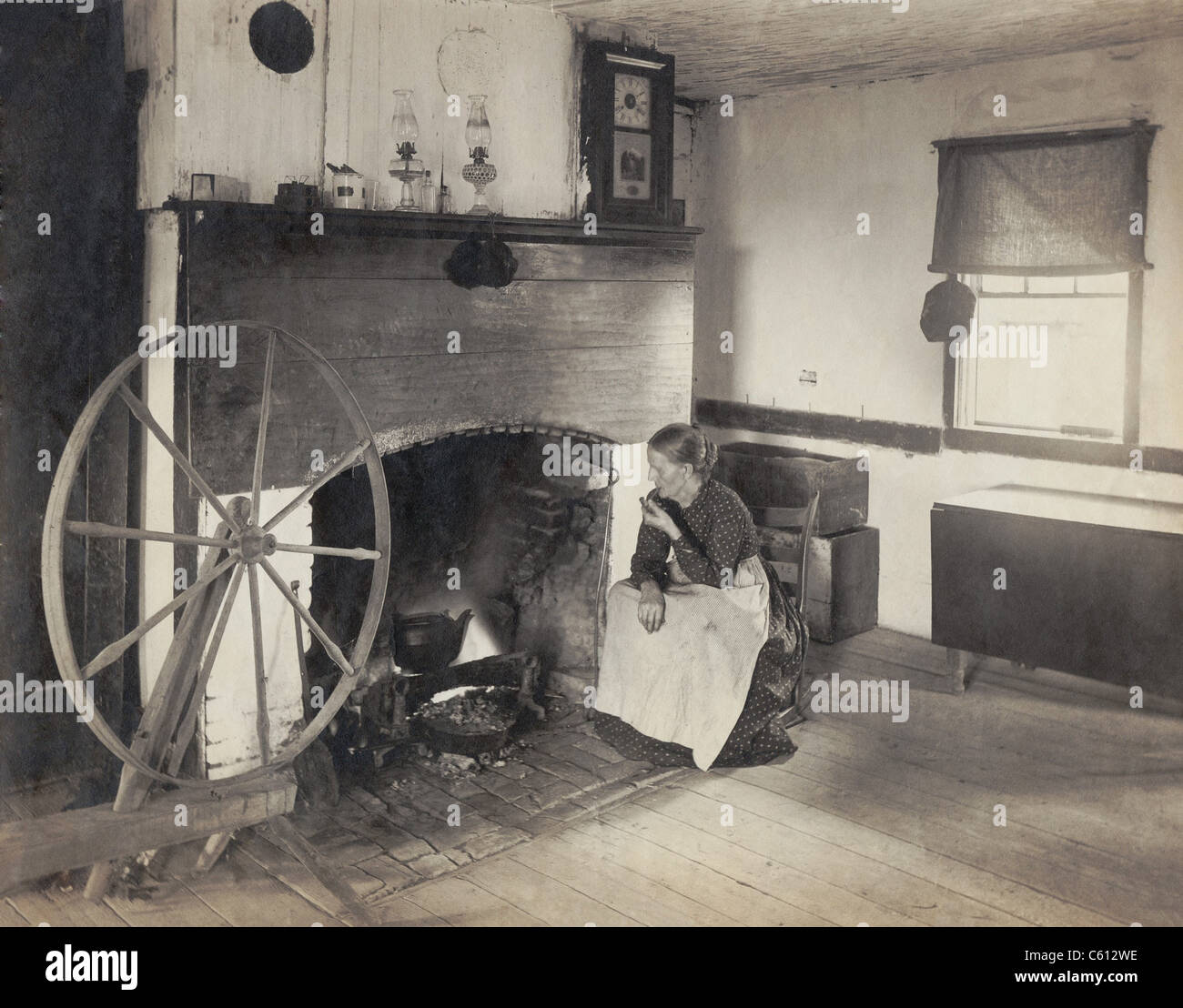 ALTEN ZEITEN. Applying Frau gerade ihre Küche von einem Schaukelstuhl durch einen Kamin, wie sie eine Pfeife raucht. Foto um 1900, Neuerstellung des Grenze Lebensstils des frühen 19. Jahrhunderts gemacht. Stockfoto