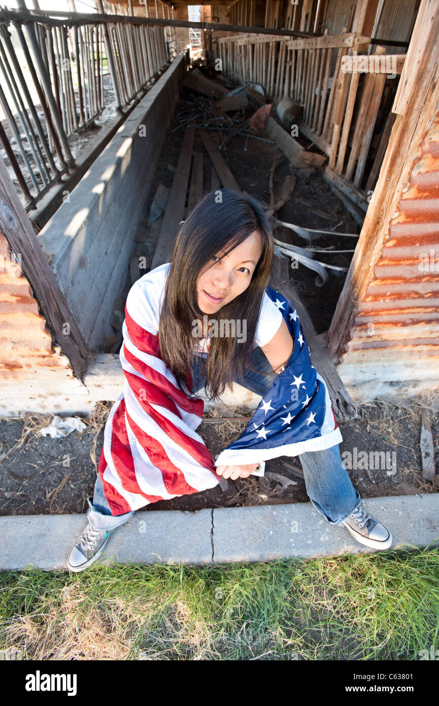 Patriotische Asiatin mit einer amerikanischen Flagge sitzt auf einer alten verlassenen Scheune Haustür Stockfoto