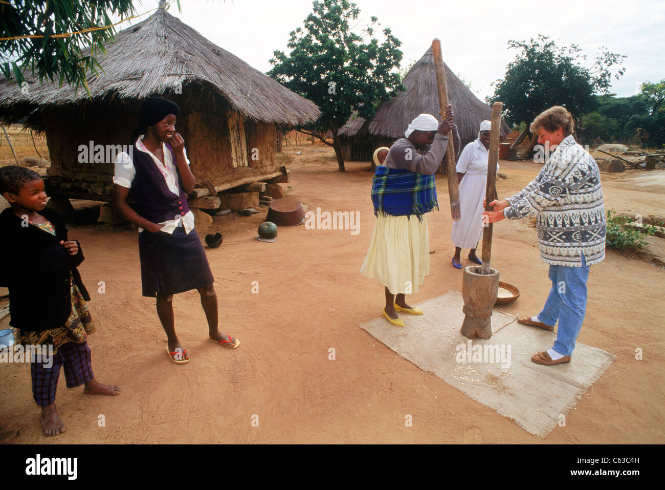 Weibliche Touristen mit Dorffrauen hämmerte Mais für Maisbrot in ländlichen Simbabwe Stockfoto