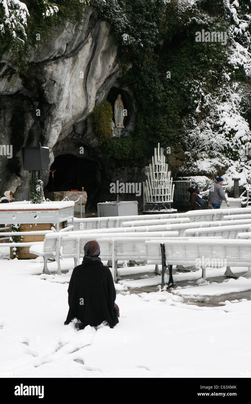 Lourdes im Winter: eine betende Pilger an der Grotte, die Wallfahrtskirche unserer lieben Frau von Lourdes. Stockfoto