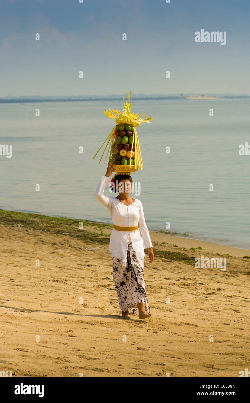 Asiatische Frau in traditioneller balinesischer Kleidung, die an einem Strand mit Obstkorb oder Canang-Sari-Opfergabe auf ihrem Kopf läuft, mit dem Indischen Ozean im Hintergrund. Stockfoto