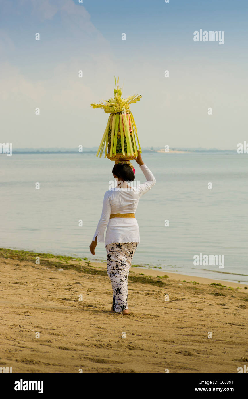 Balinesin in traditioneller Tracht, die am Strand mit Obstkorb oder Canang Sari auf dem Kopf herumläuft. Stockfoto
