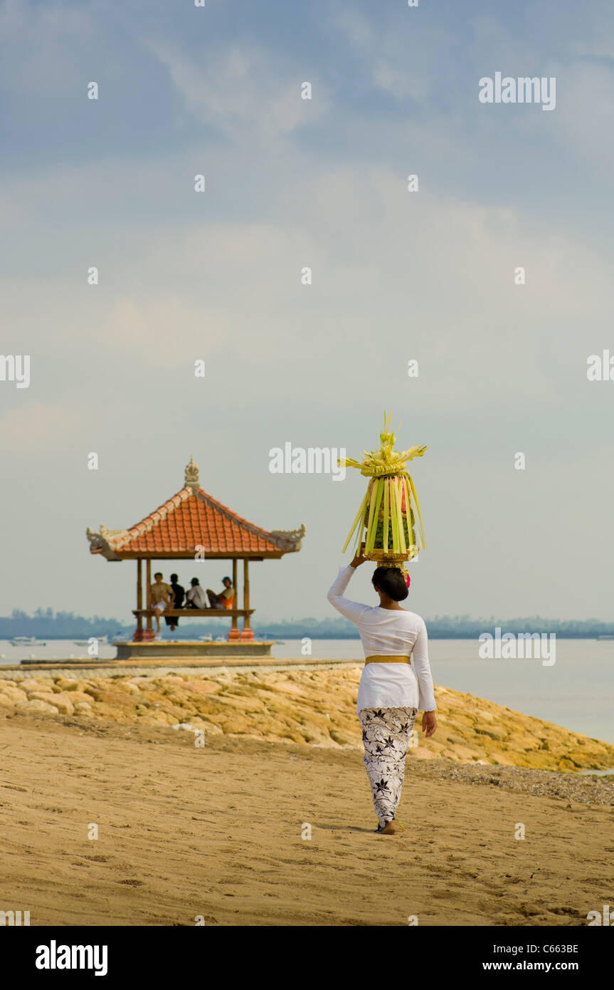 Balinesin in traditioneller Tracht, die am Strand zu einer Pergode geht und einen Korb mit Obst oder Canang Sari auf dem Kopf trägt. Stockfoto