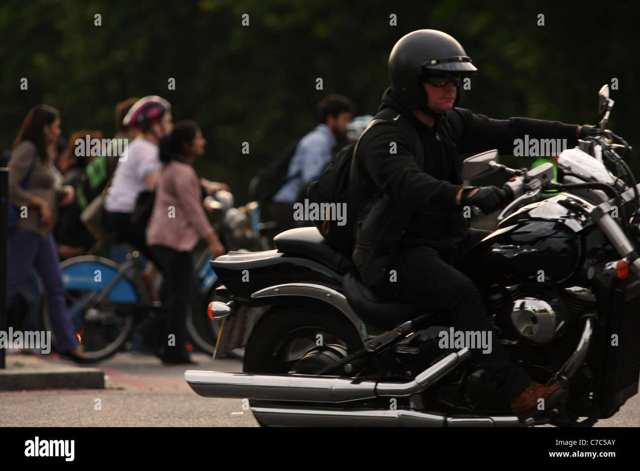 Ein Motorradfahrer Reisen rund um einen Kreisverkehr mit Fußgängern und Radfahrern, die darauf warten, eine Straße im Hintergrund Stockfoto