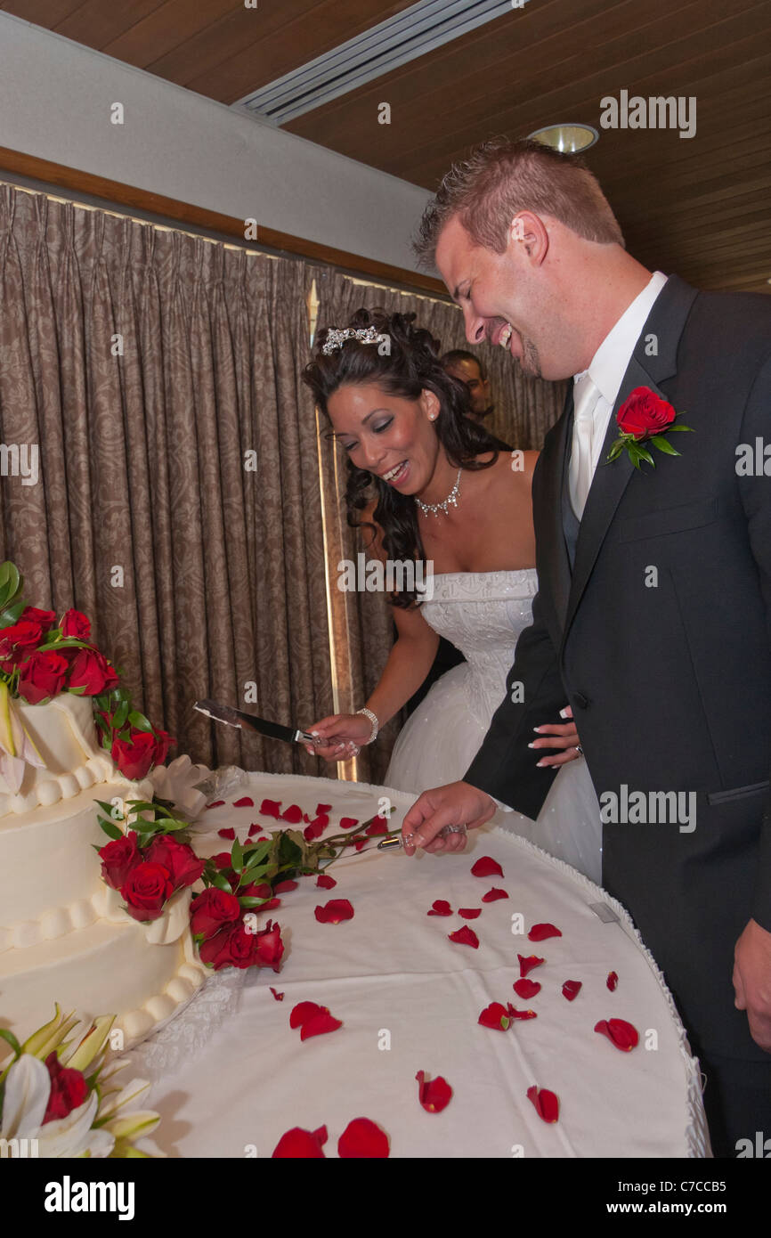 Interracial Pärchen schneiden große verziert mit roten Rosen Kuchen während der Rezeption Trauung Stockfoto