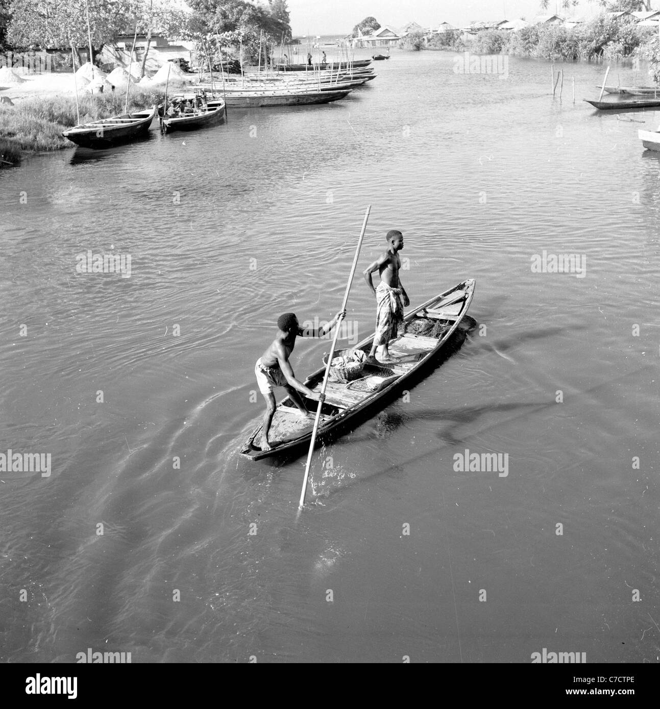In den 1950er Jahren, Nigeria, Afrika, zwei Jungen, die auf einem Kanu auf einem breiten Fluss stehen, von denen einer einen langen Pol benutzt, um das Schiff zu steuern, auf diesem Bild, das von J. Allan Cash aufgenommen wurde. Stockfoto
