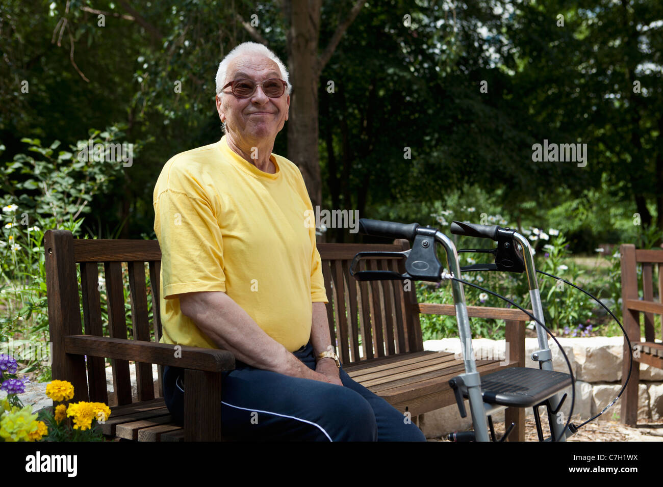 Ein senior Mann mit einer Gehhilfe ruht auf einer Bank auf dem Hof von seinem Pflegeheim Stockfoto