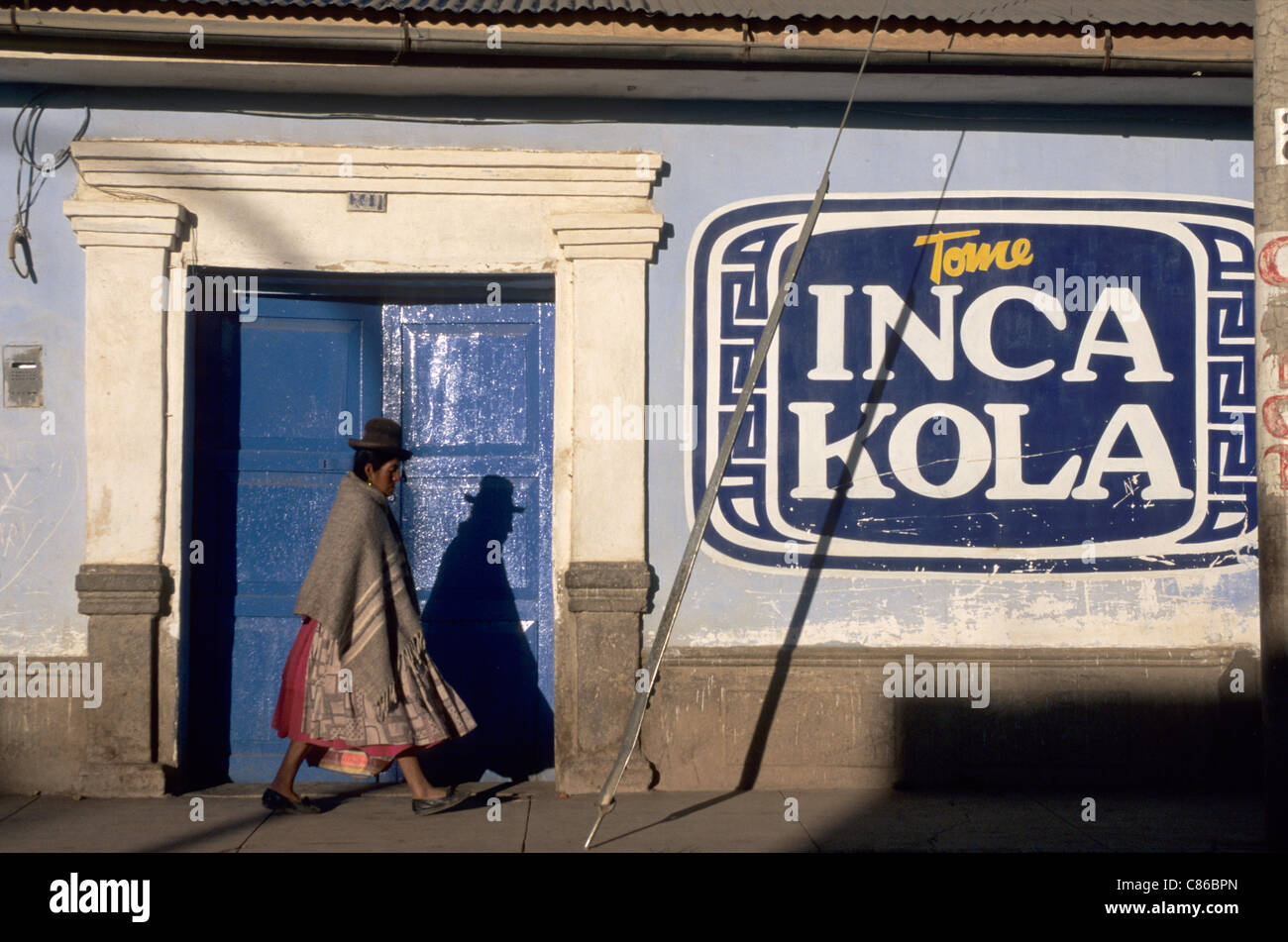 Peru. Frau in traditioneller Kleidung, vorbei an einer blauen Tür mit einem großen Inca Kola-Zeichen. Stockfoto