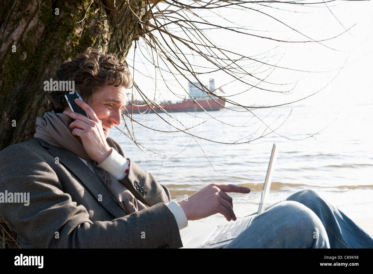 Deutschland, Hamburg, Mann mit Laptop und Handy in der Nähe von Elbe am Flussufer mit Containerschiff im Hintergrund Stockfoto