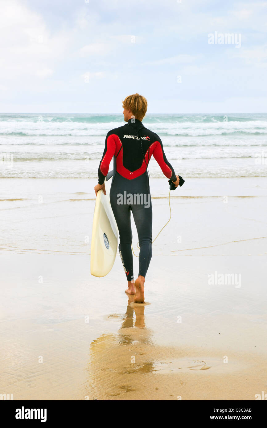 Ein Mann trägt einen Neoprenanzug zu Fuß über den Strand bis zum Meer mit einem Surfbrett in Perranporth, Cornwall, England, UK. Stockfoto