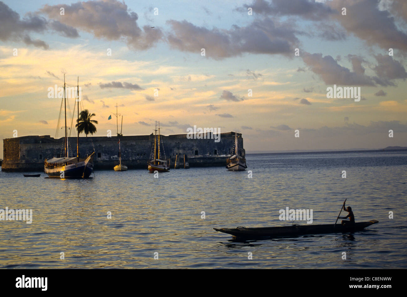 Salvador, Bahia, Brasilien. Ein Mann paddelt eine Kanu vorbei an der alten Festung Sao Marcelo, in der Abenddämmerung einzige Palme und Segelboote Stockfoto