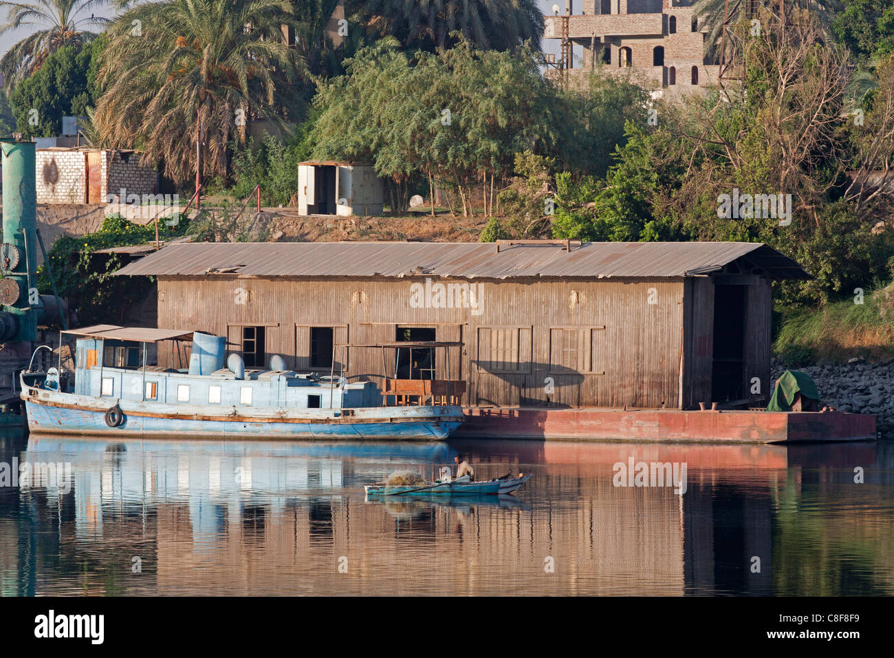 Abschnitt des Nil Flusses mit kleinen blauen, vereinzelt Spritztülle Boot ankern neben langen braunen Schuppen mit Fischer im Boot Stockfoto
