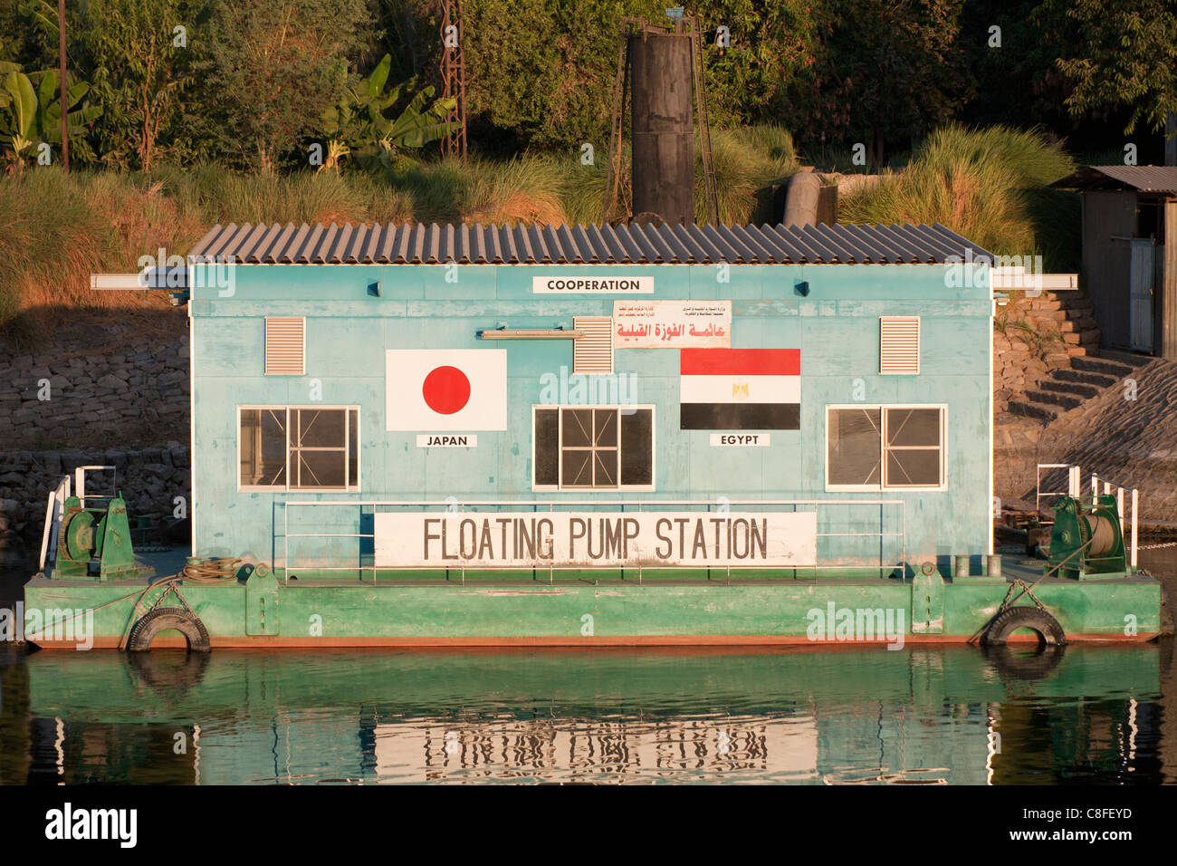 Nahaufnahme von Grün und blau, die Pumpstation am Nil-Ufer zeigt japanische und ägyptische Flaggen, spiegelt sich in dem ruhigen Wasser schweben Stockfoto