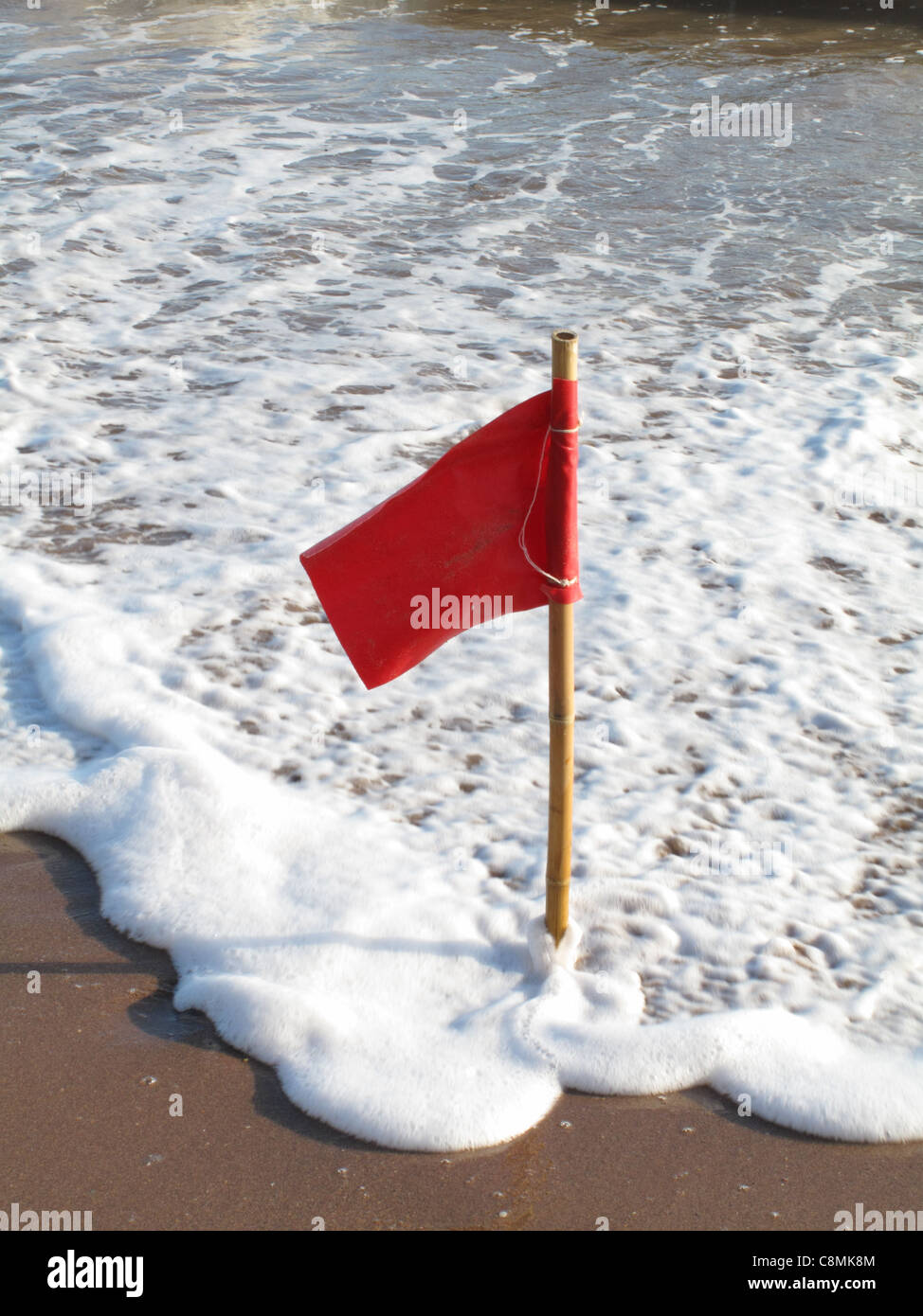 Eine rote Fahne auf einem Mast in den Sand mit der schäumenden Flut um ihn herum eingefügt. Stockfoto