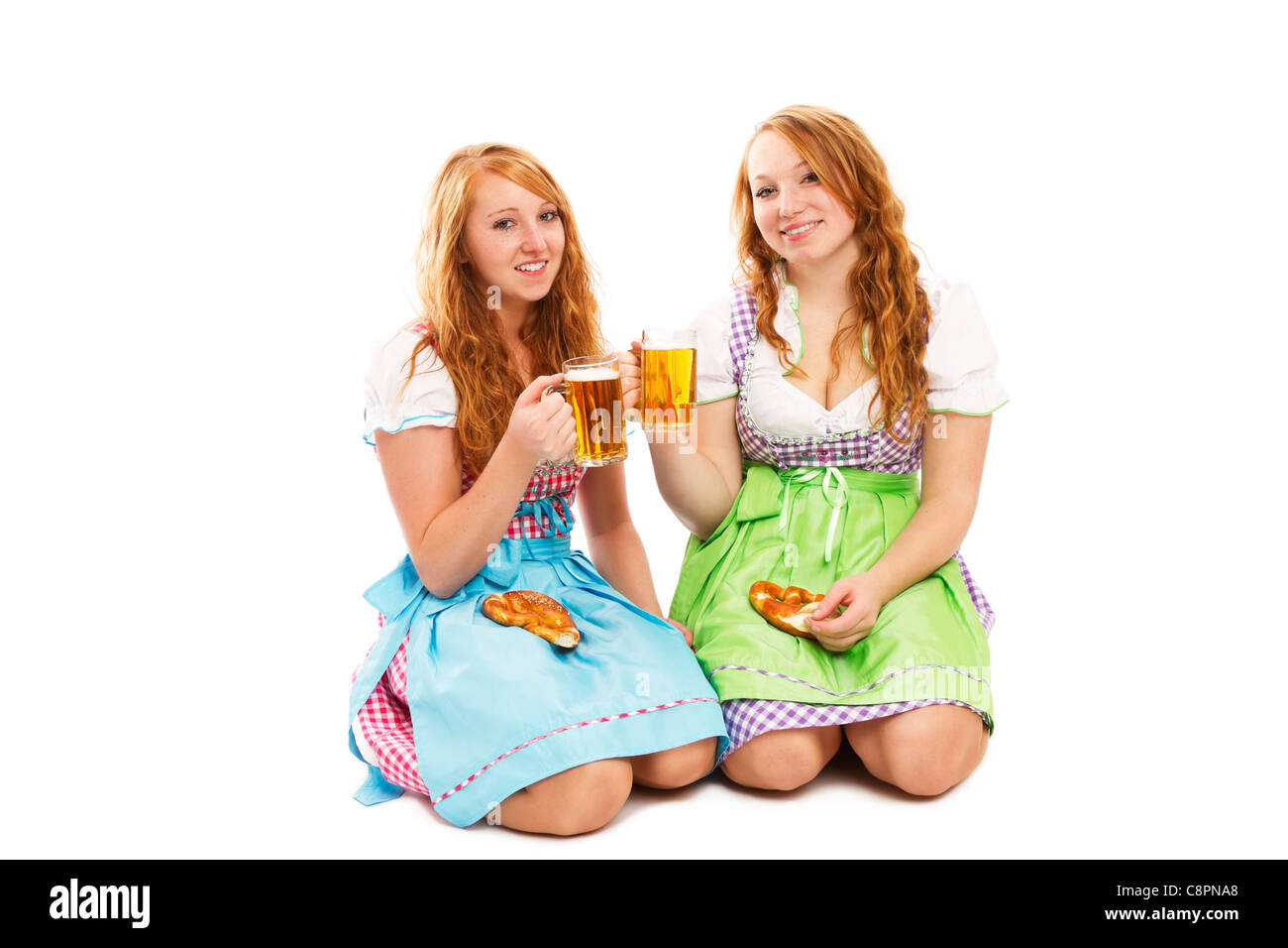 zwei bayerische Mädchen mit Brezeln und Bier kniend am Boden auf weißem Hintergrund Stockfoto