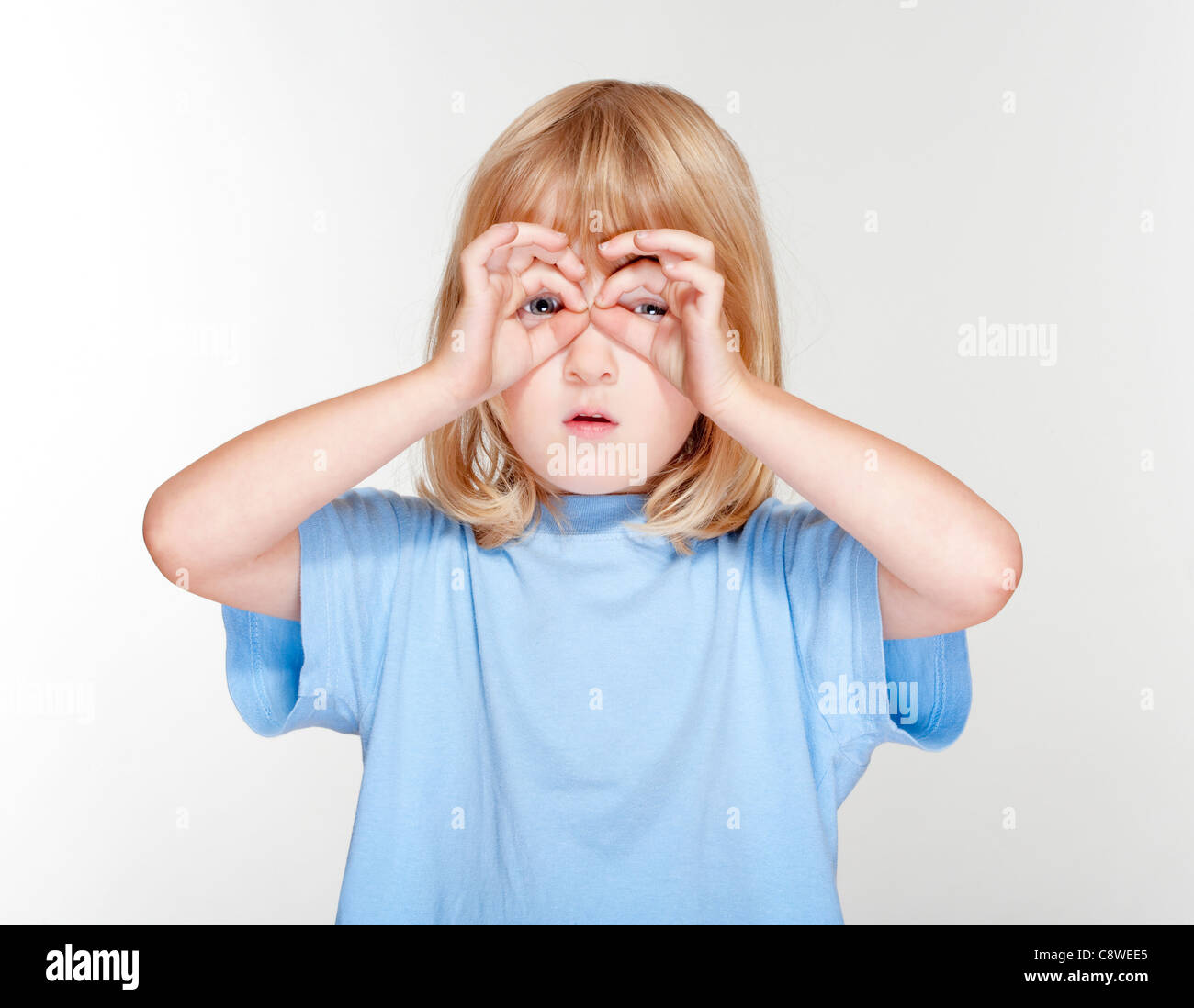 Junge mit langen blonden Haaren, die auf der Suche durch die Finger als Fernglas - isoliert auf grau Stockfoto