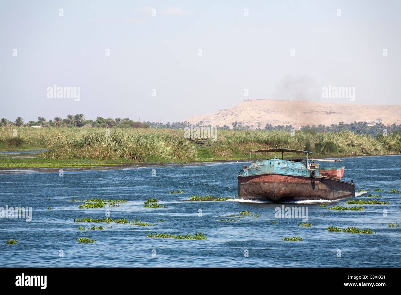 Ein Teil der Nil mit Schlepper Boot Segeln in Richtung Kamera mit Palmen und Bäumen auf dem Ufer Bergen im Hintergrund, Ägypten Stockfoto
