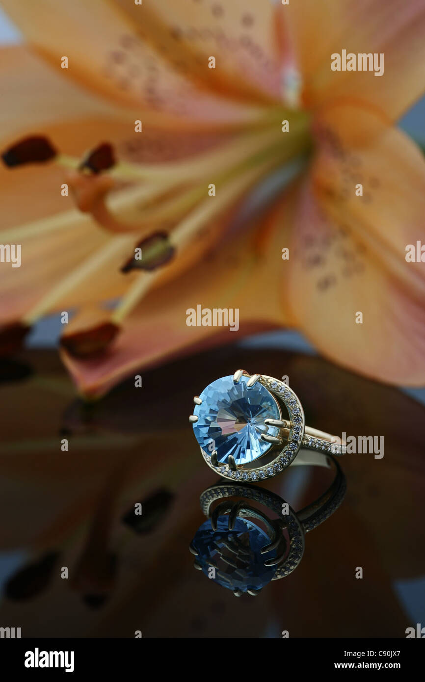 Goldener Ring mit ein großes Juwel auf der spiegelnden Oberfläche und eine große orange Lilie auf einem Hintergrund Stockfoto