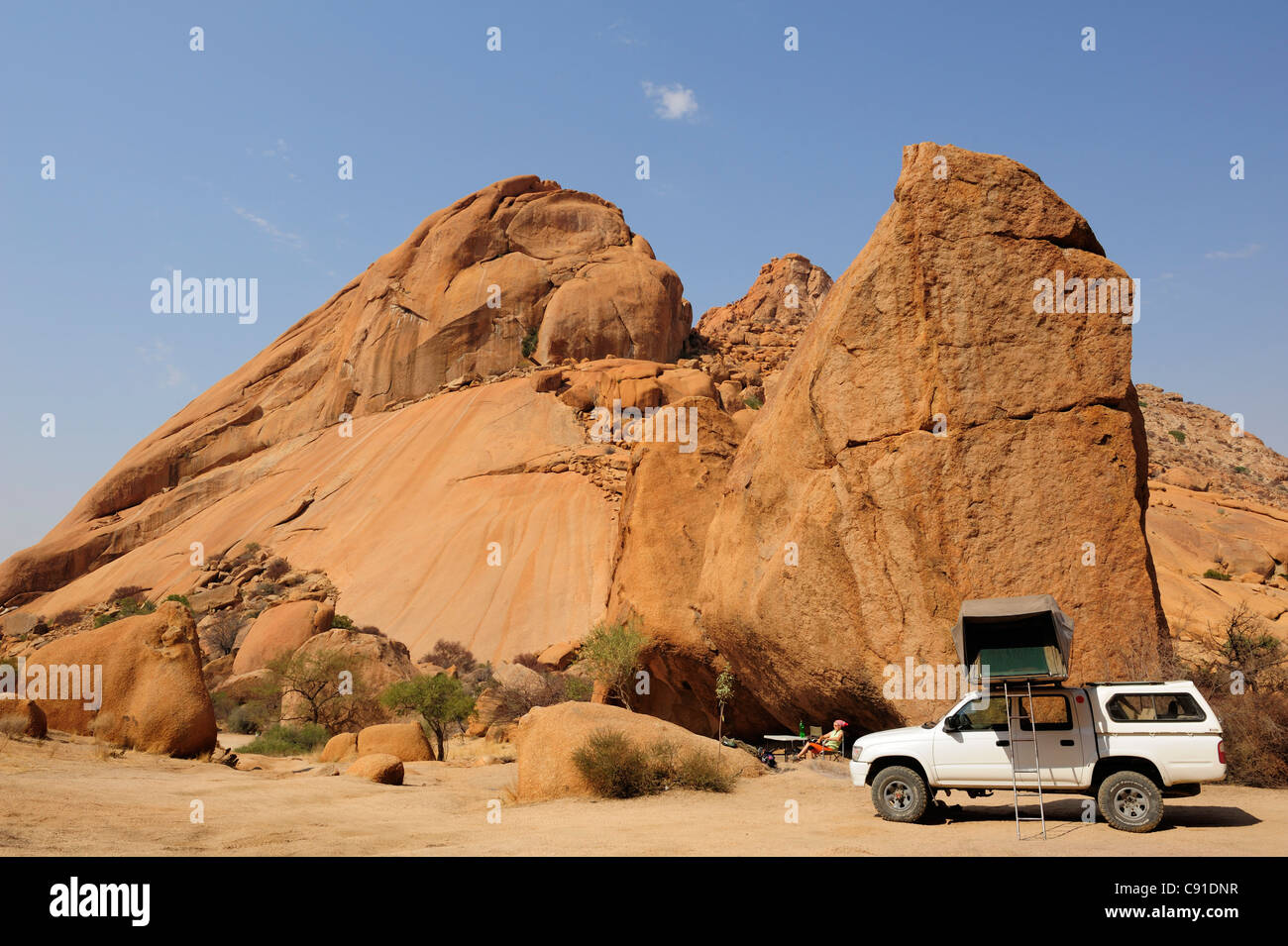 Frau sitzt vor roten Felswand, Auto mit Dachzelt im Vordergrund, große Spitzkoppe, Namibia Stockfoto