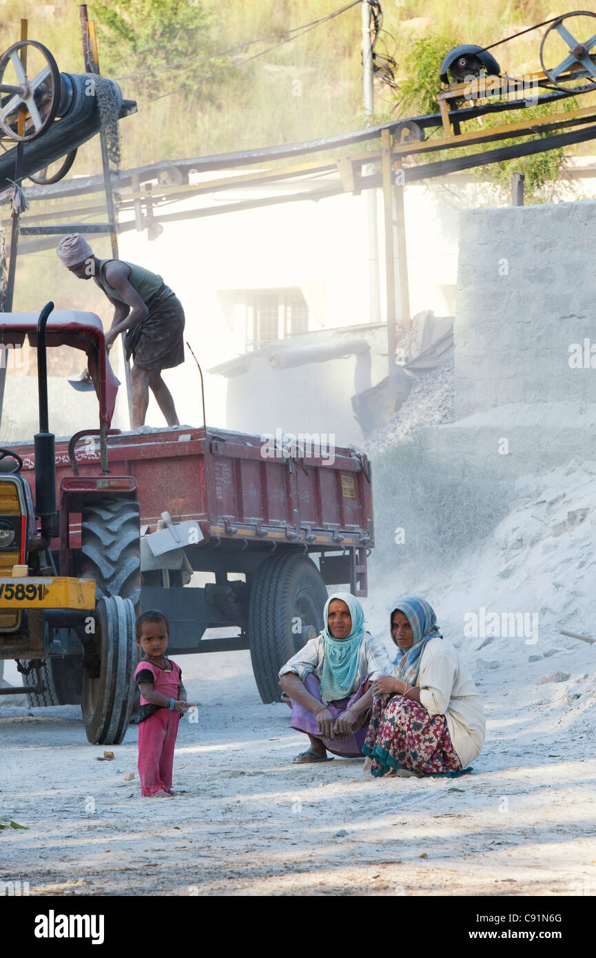 Indische Frauen und Kind sitzen neben dem Traktor und Anhänger, umgeben von Staub, ungeschützt, an einem Stein Quetschungen funktioniert. Andhra Pradesh, Indien Stockfoto