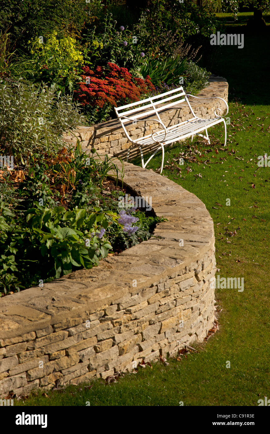 Gebogene Cotswold Steinmauer mit erhöhten Blumenbeet im englischen Garten und Weißmetall Bank im Herbst. Stockfoto
