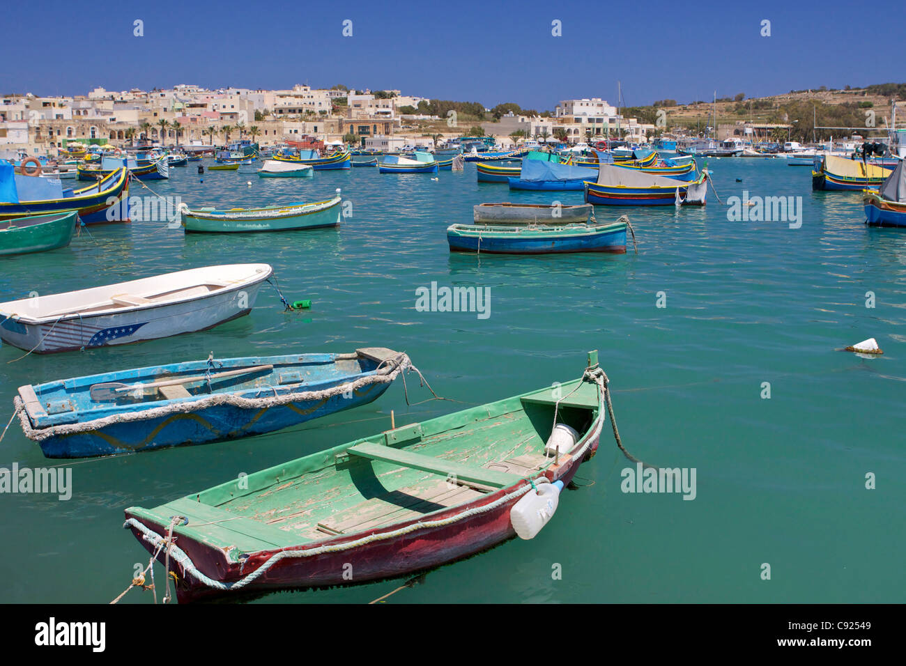 Ruderboote und Luzzu - traditionellen maltesischen Fischerboot vor Anker im Hafen von Marsaxlokk mit der Stadt im Hintergrund. Stockfoto