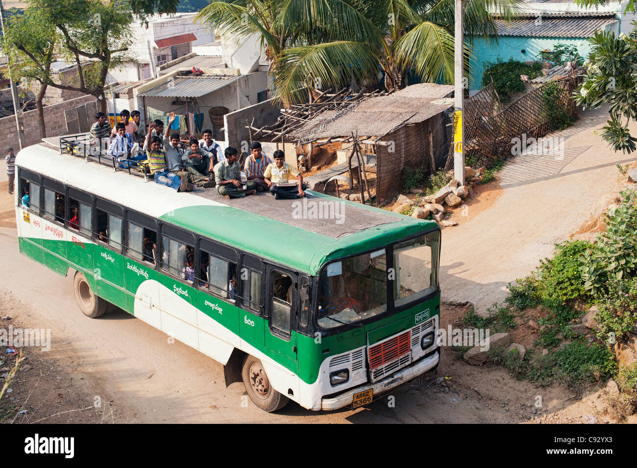 Indische Bus entlang einer ländlichen indischen Dorf Straße unterwegs mit Jungs im Teenageralter sitzen auf dem Dach. Andhra Pradesh, Indien Stockfoto