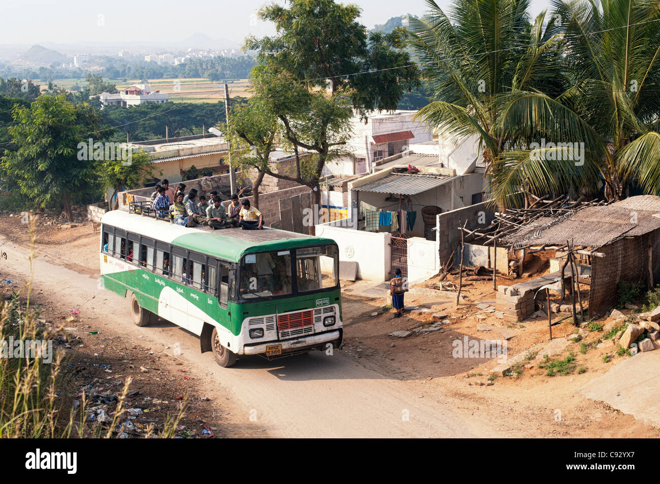 Indische Bus entlang einer ländlichen indischen Dorf Straße unterwegs mit Jungs im Teenageralter sitzen auf dem Dach. Andhra Pradesh, Indien Stockfoto