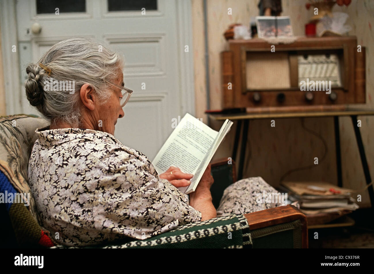 Alte Dame Buch sitzt in einem Sessel mit einem alten Radio im Hintergrund festlegen Stockfoto