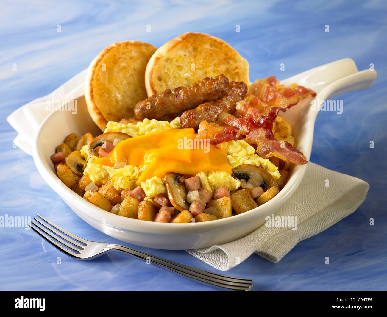 Eine Frühstück-Pfanne aus Kartoffeln, Pilze und Zwiebeln bedeckt mit Eiern und Käse und serviert mit Speck, Wurst und muffins Stockfoto
