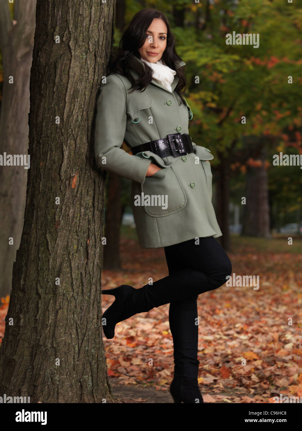 Modisch gekleidet für die Herbstsaison, schöne lächelnde junge Frau trägt einen grünen Mantel, lehnte sich gegen einen Ahornbaum im park Stockfoto