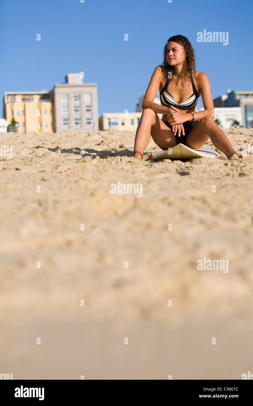 Junge Frau sitzt neben Surfbrett am Strand.  Bondi Beach, Sydney, New South Wales, Australien Stockfoto