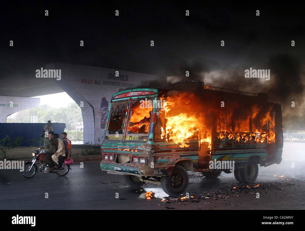 Motorradfahrer fahren durch ein brennendes Fahrzeug, das in Flammen von unbekannten Schurken bei Gewalt in Karachi auf Dienstag, 27. März 2012 festgelegt wurde. Stockfoto