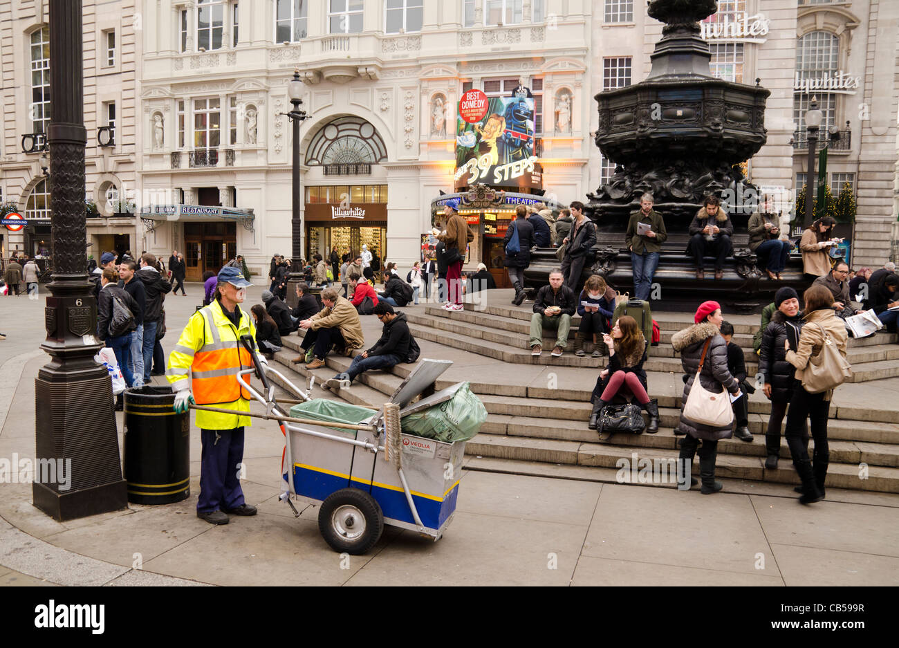 Eine Straße sauberer und seinen Karren vor Menschen sitzen auf den Stufen der Eros-Statue Piccadilly Circus London UK Stockfoto